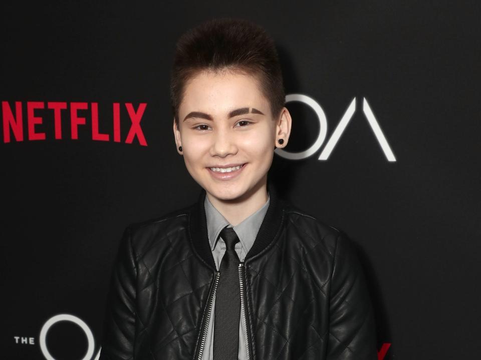 Ian smiling with short hair and earrings and a suit and bomber jacket.