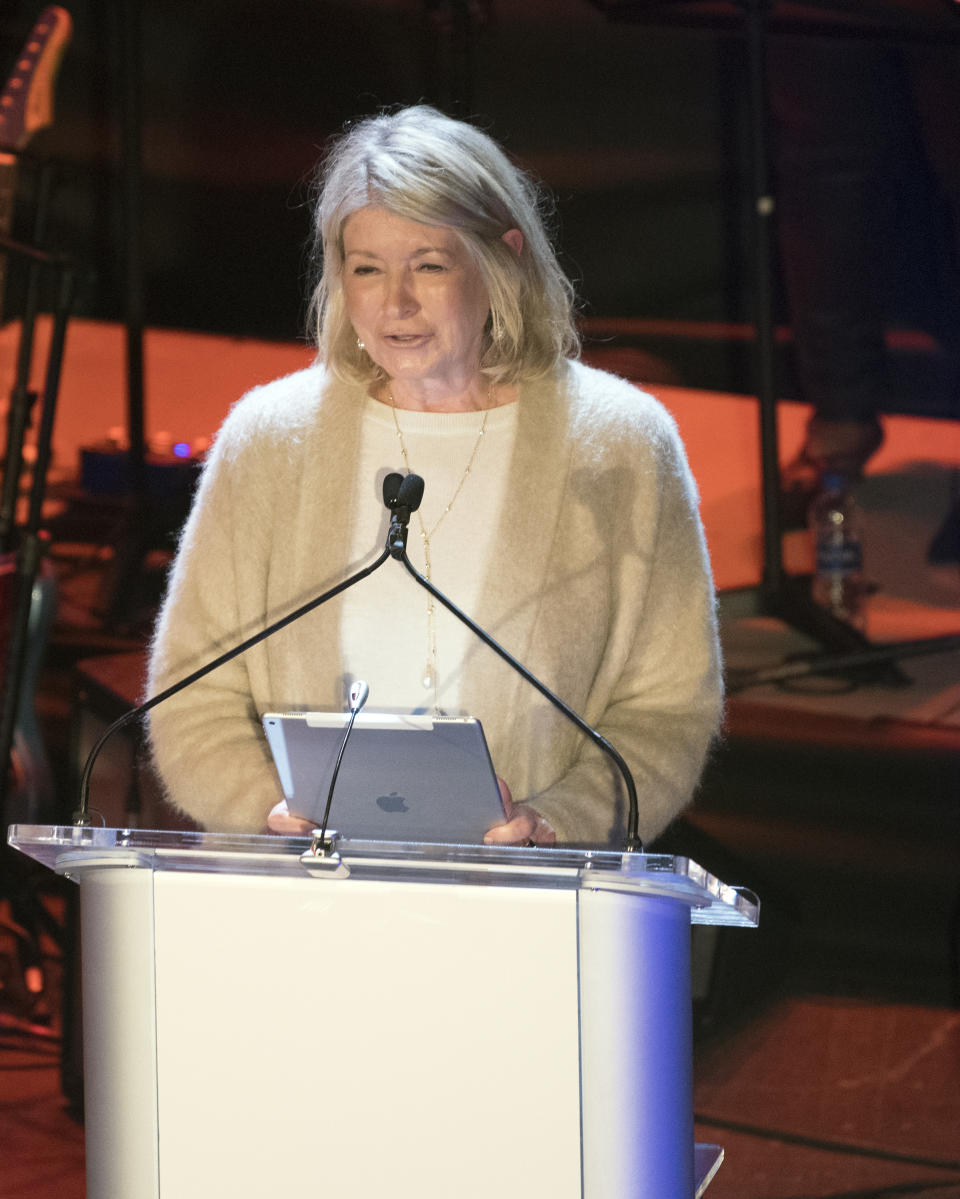 Martha Stewart stands at a podium holding an iPad, wearing a soft, casual outfit, speaking at an event