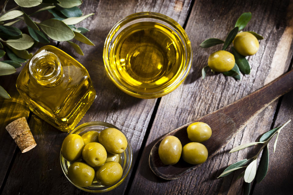 Top view of an olive oil bottle and a little glass bowl filled with green olives on rustic wood table. Two olives with leaves are at the top-right while a bowl filled with olive oil is at the center-top beside the two olives. An olive tree branch is at the left-top corner. A wooden spoon with three olives comes from the right. Predominant colors are gold, green and brown. DSRL studio photo taken with Canon EOS 5D Mk II and Canon EF 100mm f/2.8L Macro IS USM (Photo: fcafotodigital via Getty Images)