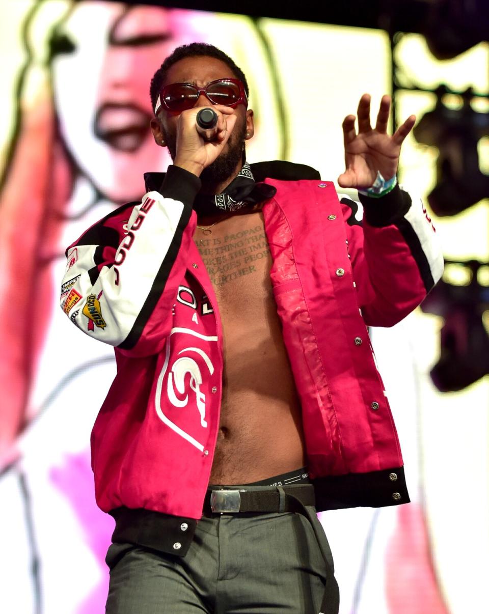 An R&B singer in a jacket and no shirt performs onstage outdoors