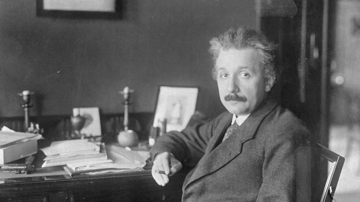 albert einstein sitting at a desk, wearing a suit and tie, looking directly at the camera