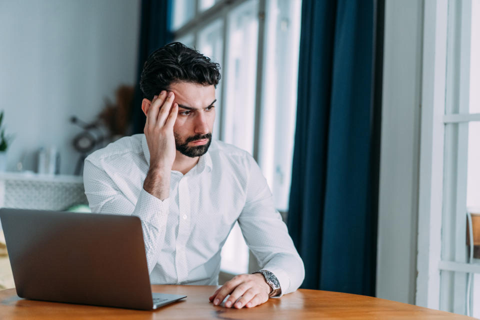 A man in a white shirt sits at a wooden table with a laptop, resting his head on his hand, looking concerned