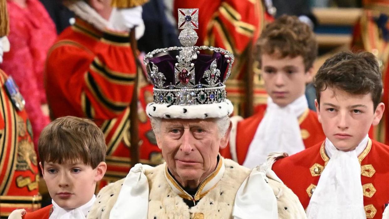 king charles walks down on an aisle with three boys following, he smiles slightly and wears a purple and bejeweled crown with a cream fur robe