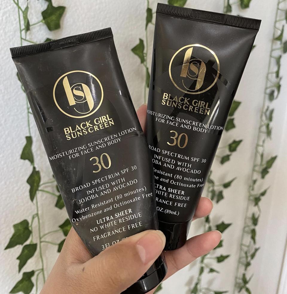 The writer holds two black tubes with gold text saying "Black Girl Sunscreen" and "30" on them