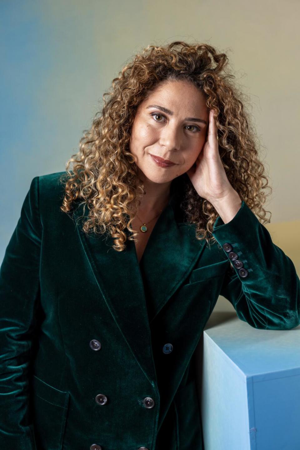 Director Mahalia Belo wears a dark jacket while smiling and leaning an arm on a cube.