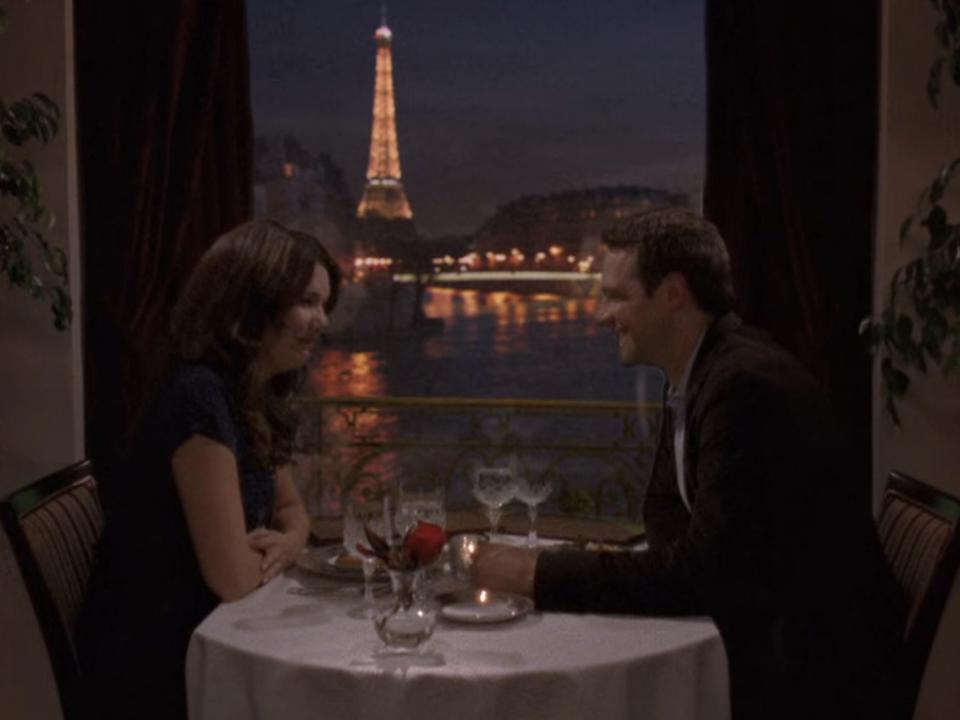 lorelai and christopher sitting at a restaurant with a view of the eiffel tower in paris