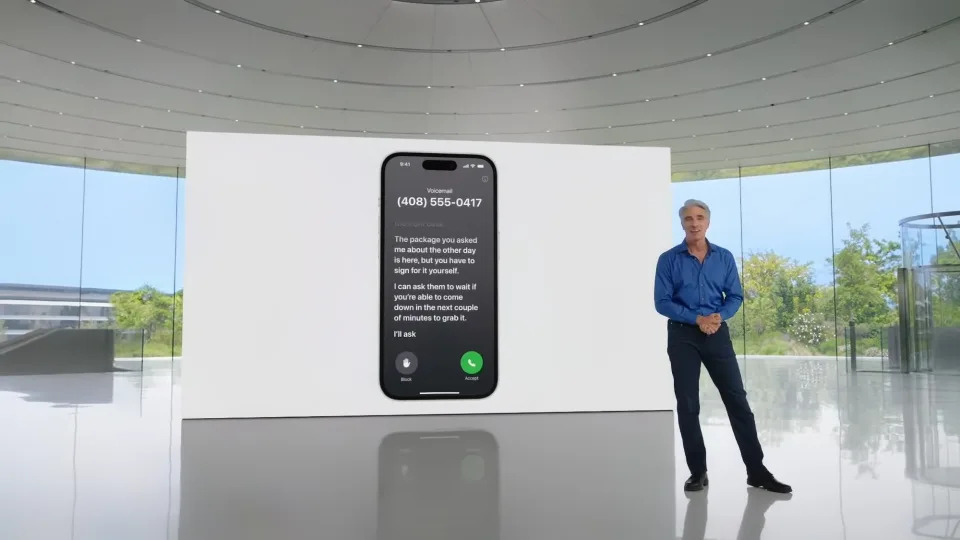 Apple executive Craig Federighi (blue button-down shirt, black slacks) standing in a room in Apple Park (modern gray aesthetic with trees visible outside the windows). On a screen behind him is a demonstration of live voicemail.