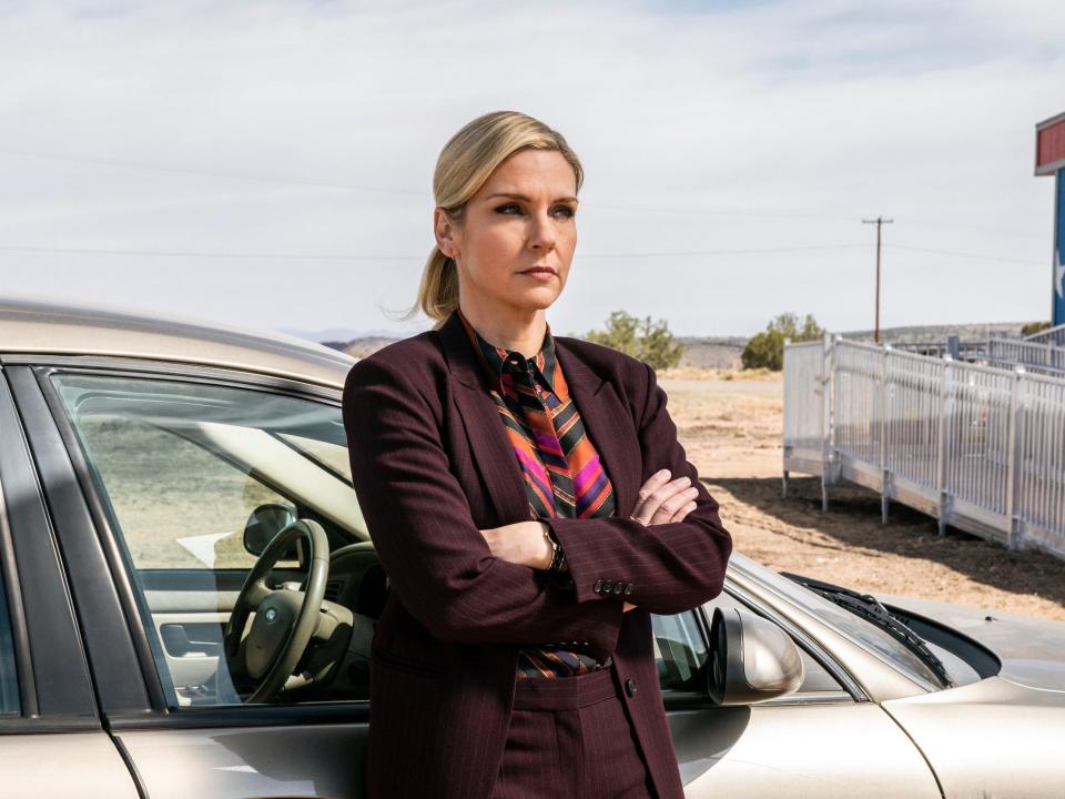 A blonde woman wearing a dark burgundy suit leans against a silver car outside a desert.