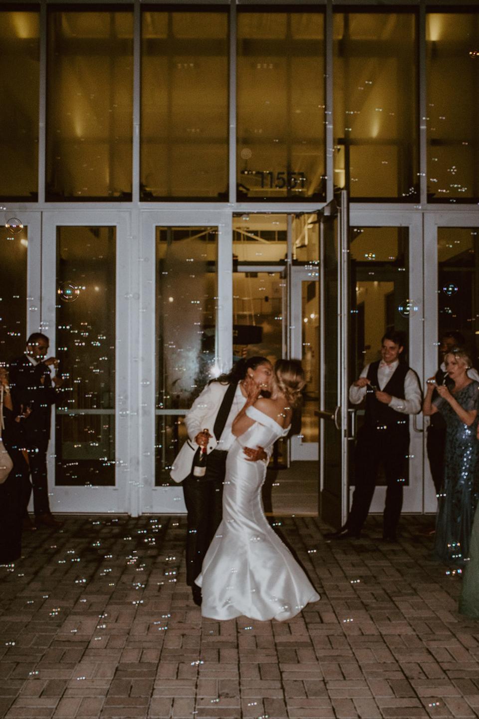 Two brides kiss as their wedding guests blow bubbles around them. One bride holds a bottle of champagne.