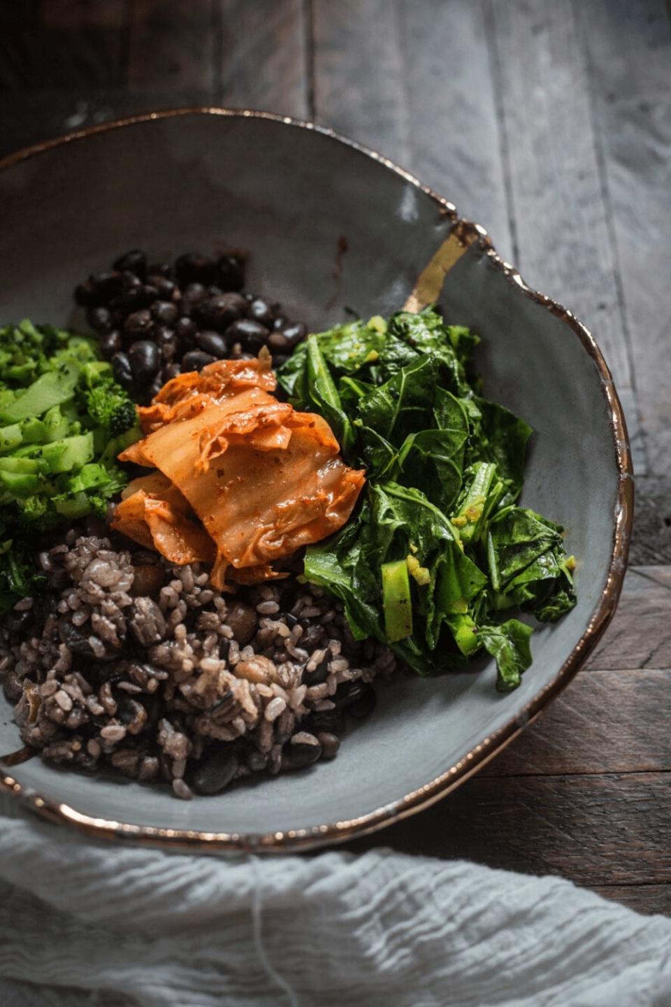 Gray ceramic bowl filled with equal parts purple rice, collard greens, kimchi, and broccoli slices.