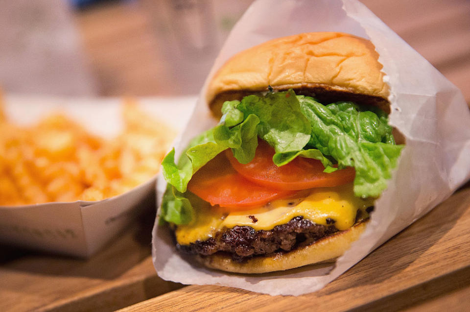 A close-up of a cheeseburger with lettuce and tomato in a wrapper, next to a container of fries