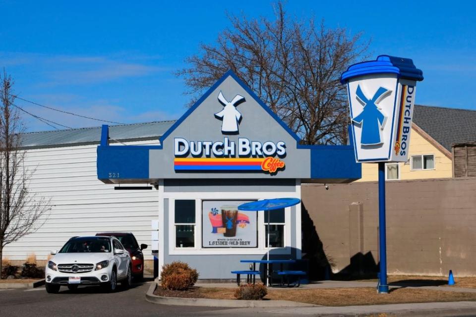 A small Dutch Bros location sits against a bright blue sky, while two vehicles are going through the company's drive-thru lane.