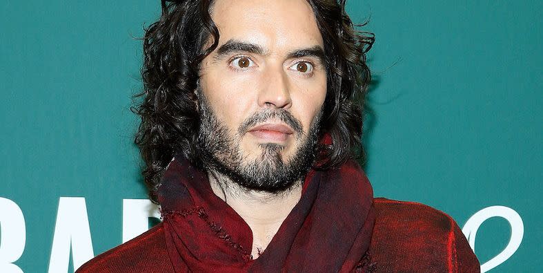 russell brand poses in a red top against a turquoise background at an event for his book launch