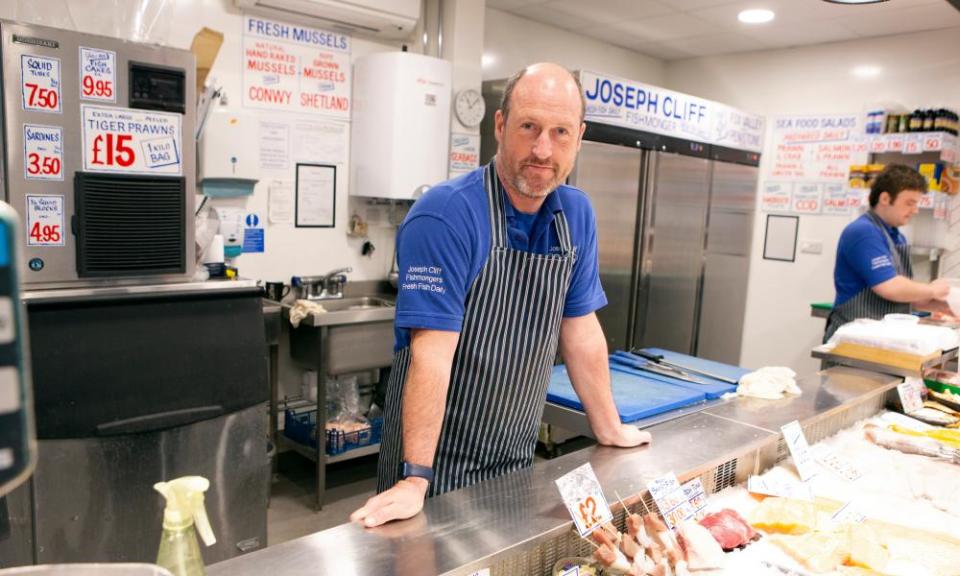 Angus Mckinlay resting both hands on his fishmonger's counter in a blue striped apron, looking at the camera