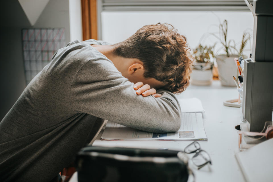 A person with curly hair rests their head on their arms on a desk cluttered with stationery and plants