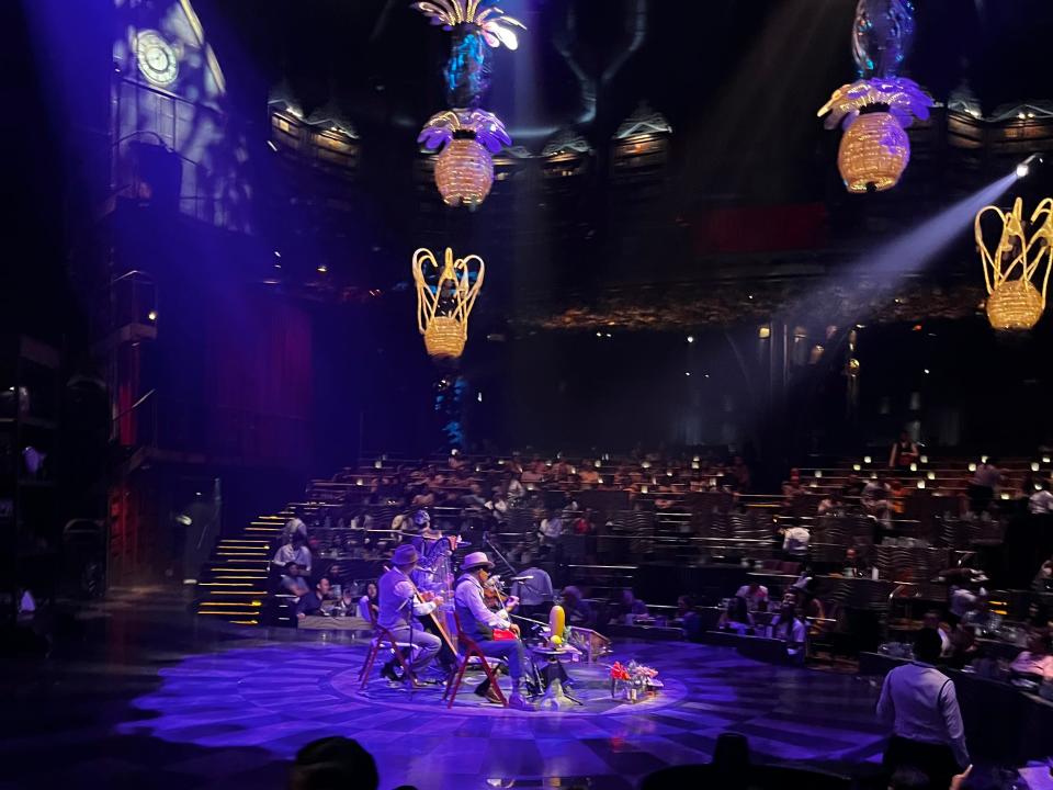 Cirque du Soleil performers play instruments in the middle of the stage and lights shaped like pineapples and other yellow lights shine overhead