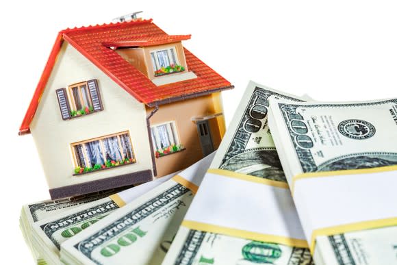 A small model of a house, perched on some stacks of hundred dollar bills