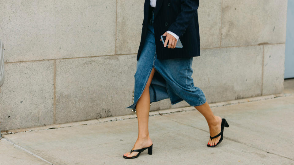 A woman walking black leather heeled flip-flops, denim skirt, and blazer