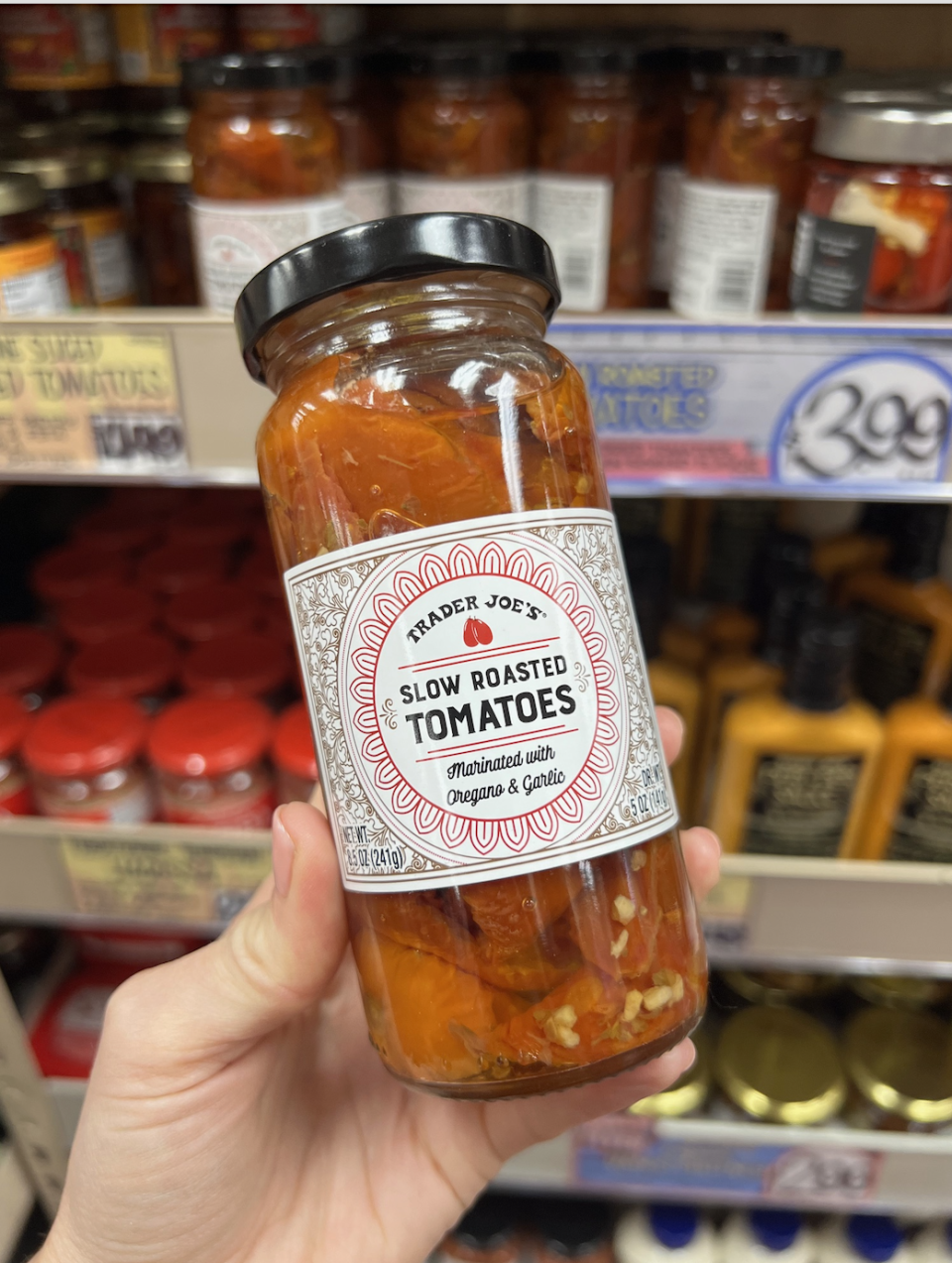 Hand holds a jar of Trader Joe's Slow Roasted Tomatoes with seasoning. Shelves of similar jars are in the background