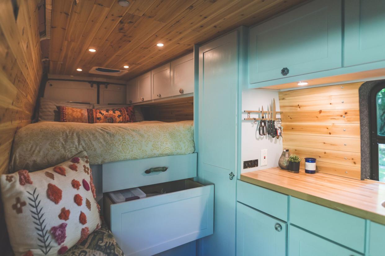 Interior of a van that a young couple live in. The shot is focused on the kitchen counter, pull-out drawers, and bed. Ther is wood paneling on the sides and roof.