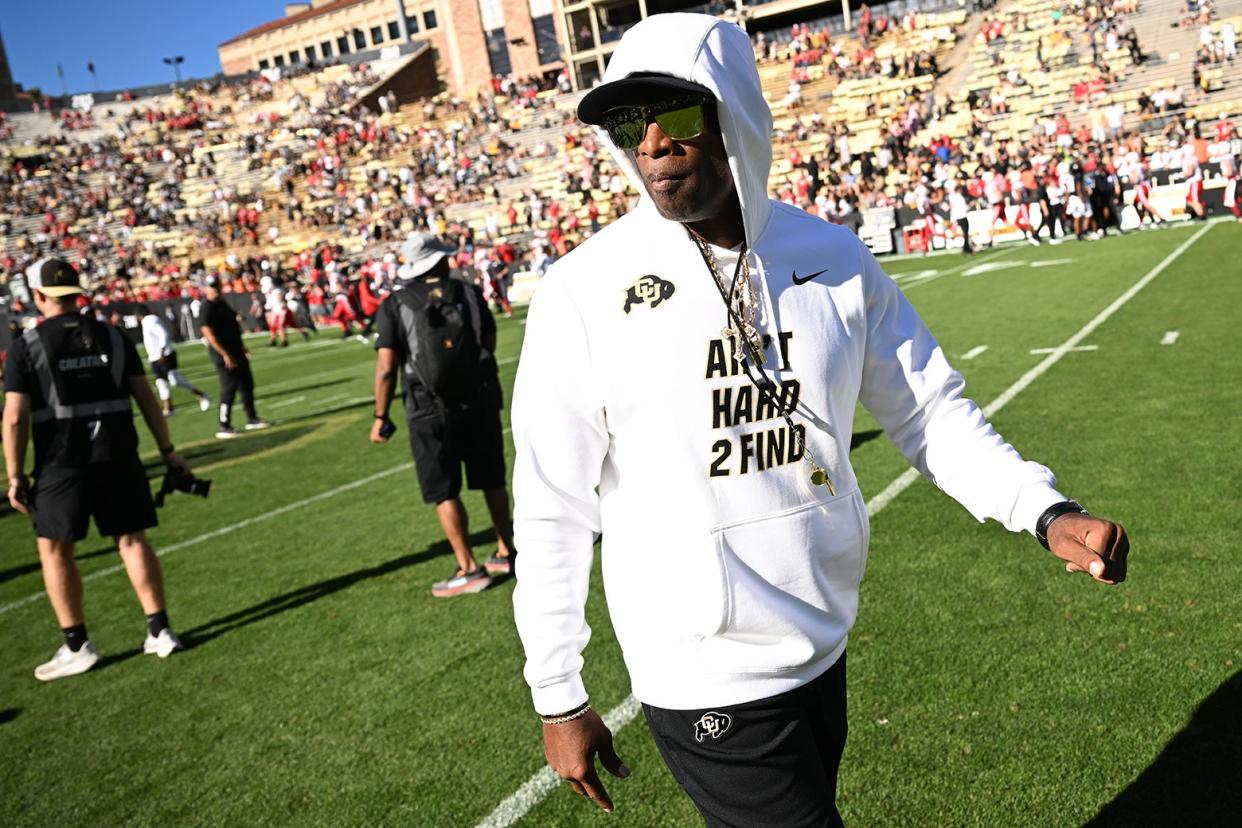 Deion Sanders in a CU hoodie that reads "Ain't Hard 2 Find" with the hood up and sunglasses on walking on the football field with crew and fans in the background.