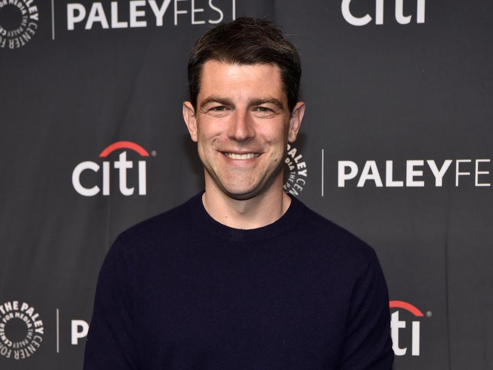 Max Greenfield in a dark blue shirt and smiling with black background and "Citi" text