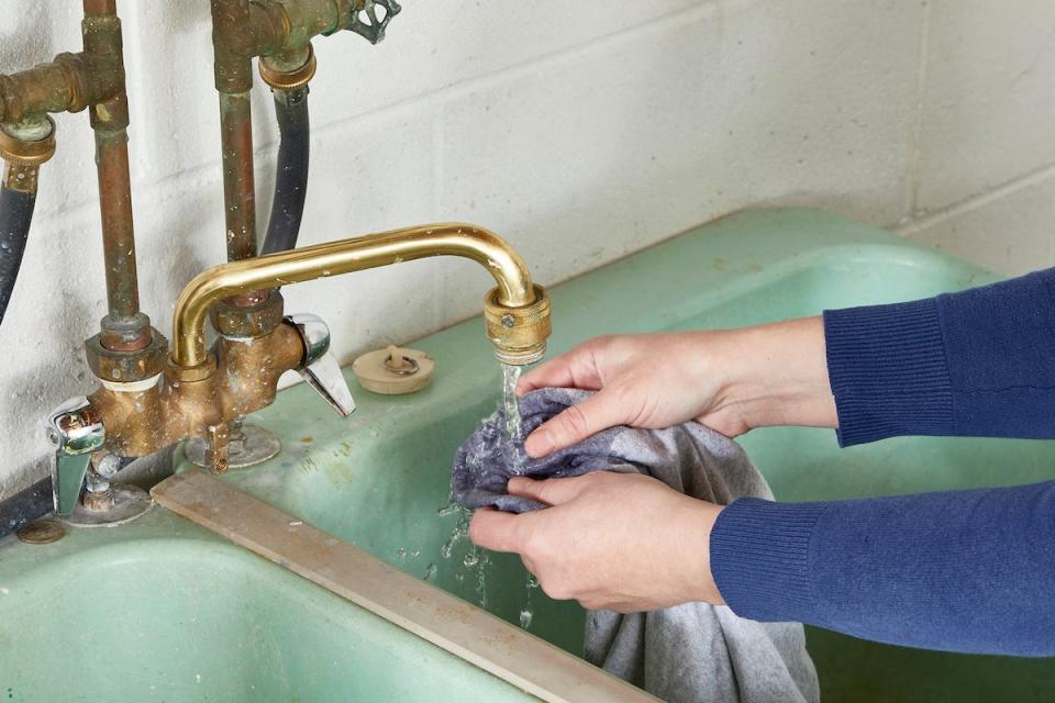 Woman rinses caulk out of a grey shirt under a sink faucet.