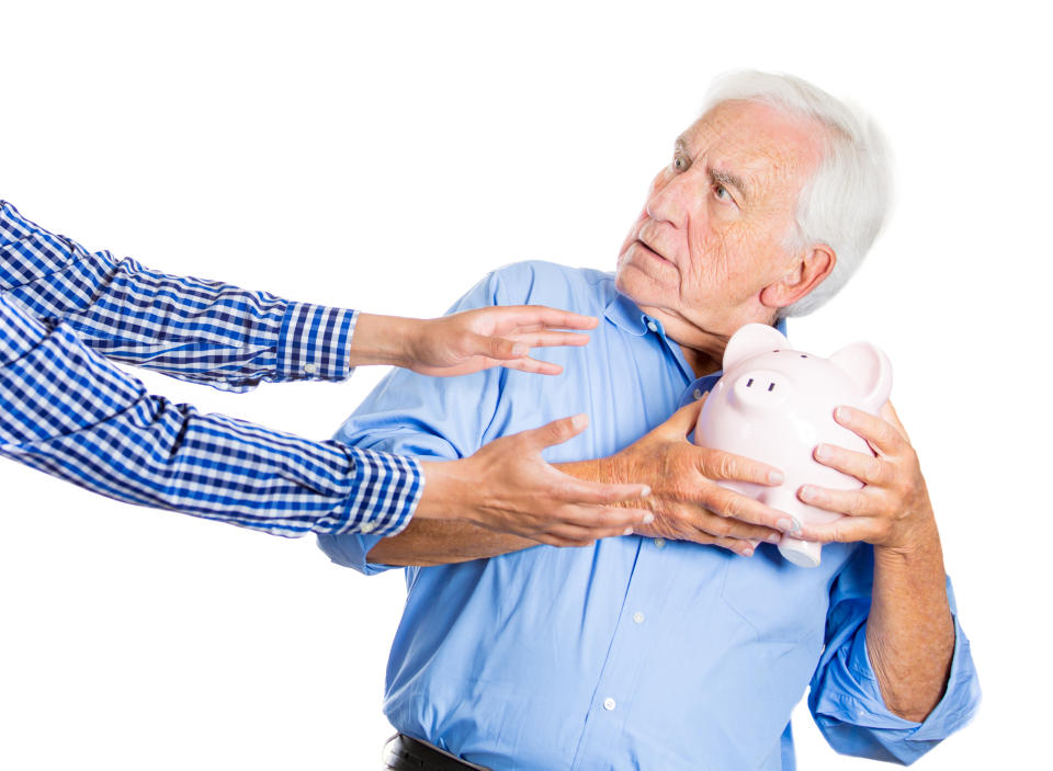 A visibly surprised senior tightly grasping his piggy bank as outstretched hands reach for it.