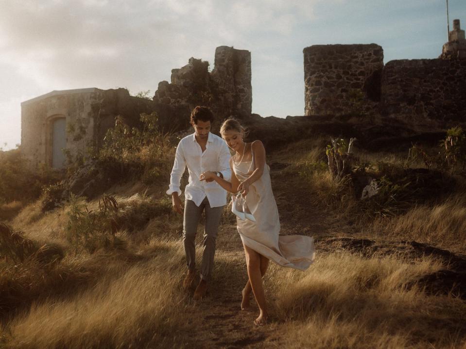 A couple hold hands and walk down a hill in front of a castle.