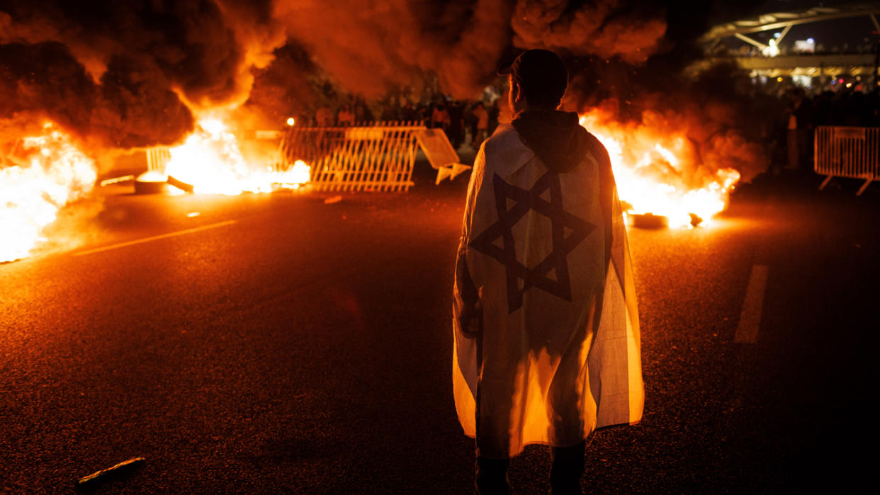 A demonstrator wears an Israeli national flag as he stands on a highway near several bonfires.