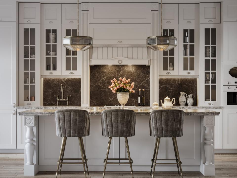 A kitchen island with intricate molding detailing along the sides, a stone countertop, three barstools, and a traditional-looking cream cabinet and stovetop area