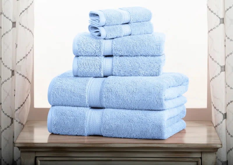 The towel set in the color blue, folded and stacked on a wooden surface with a curtained window in the background