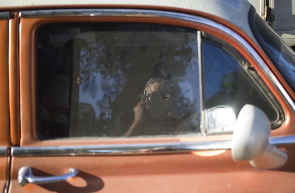 Associated Press photojournalist Desmond Boylan takes a picture of his reflection in a car window as he talks on his cell phone at a market where he was taking pictures of shoppers buying last minute items for their traditional news years dinners at a market in Havana, Cuba, Saturday, Dec. 29, 2018. Boylan, a photographer who covered war and conflict across the world before dedicating his life to documenting the joys and tribulations of daily life in Cuba for The Associated Press, died suddenly on Saturday afternoon, Dec. 29, 2018, in Havana. He was 54. (AP Photo/Desmond Boylan)