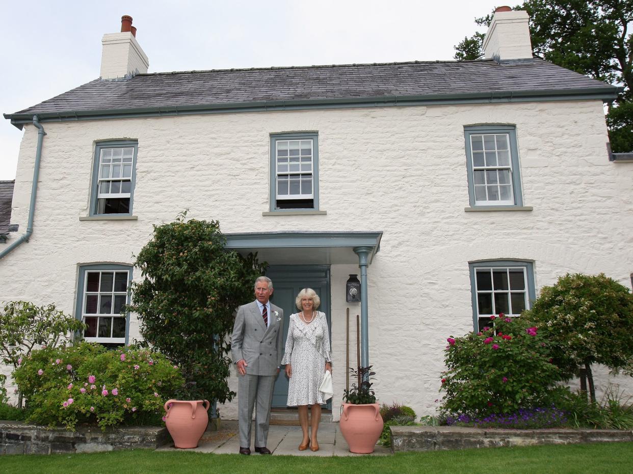 Charles and Camilla standing on the lawn in front of a small two-story cottage with white brick and a slate roof.