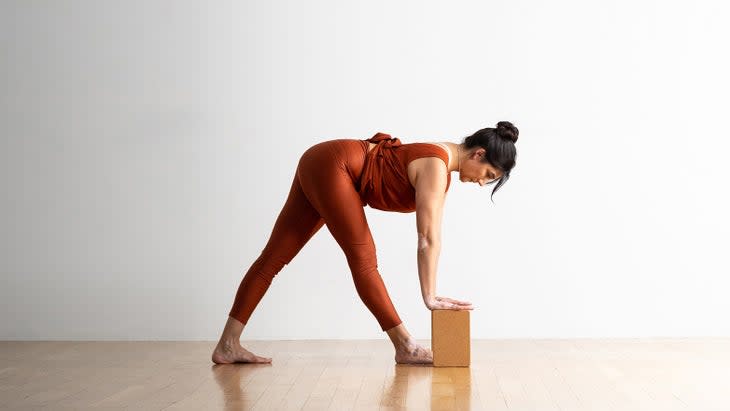 A woman with dark hair and shiny, dark orange tights and top, bends into Pyramid Pose. She places her handds on cork blocks in front of her.
