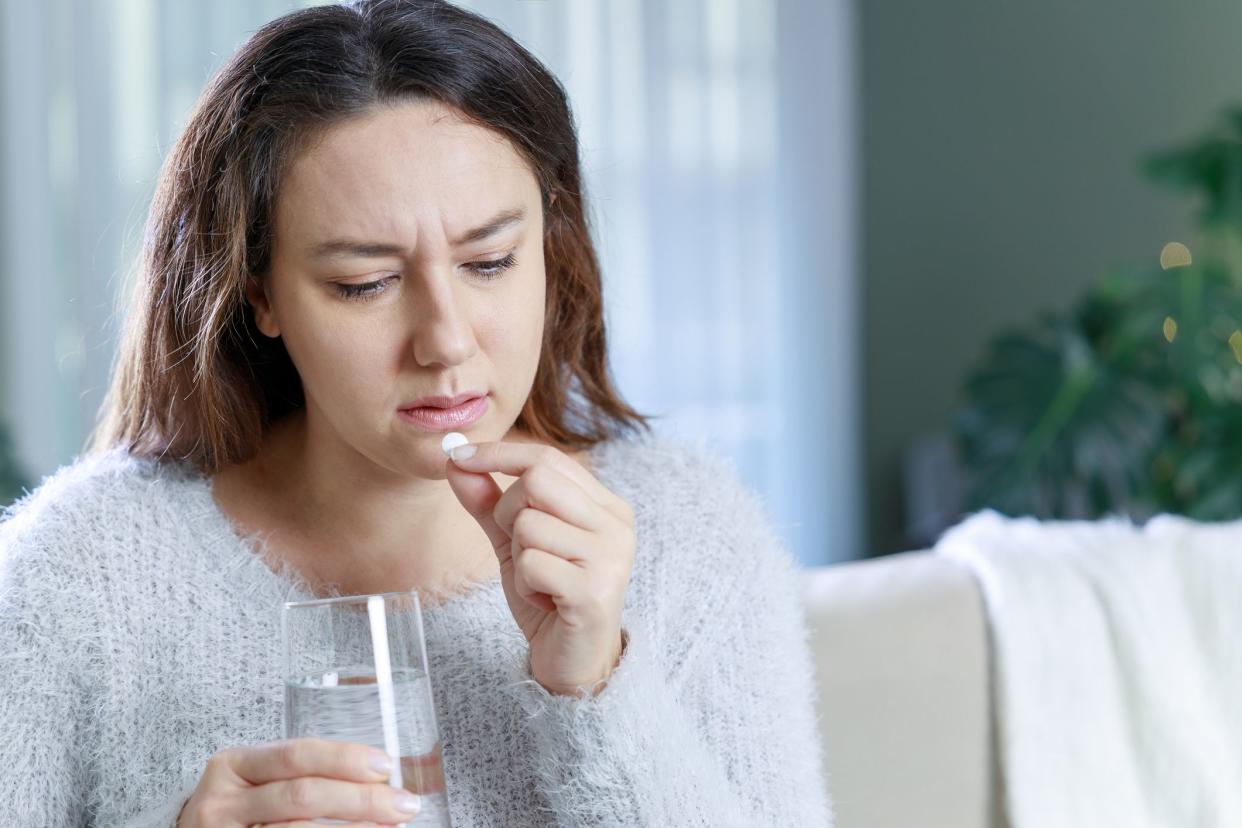 Sick woman taking pills holding a glass of water sitting on a sofa at home