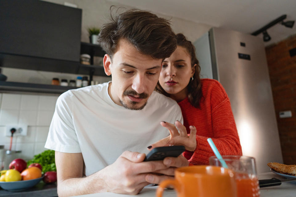 A woman looks through her boyfriend's phone while the two stand in their kitchen