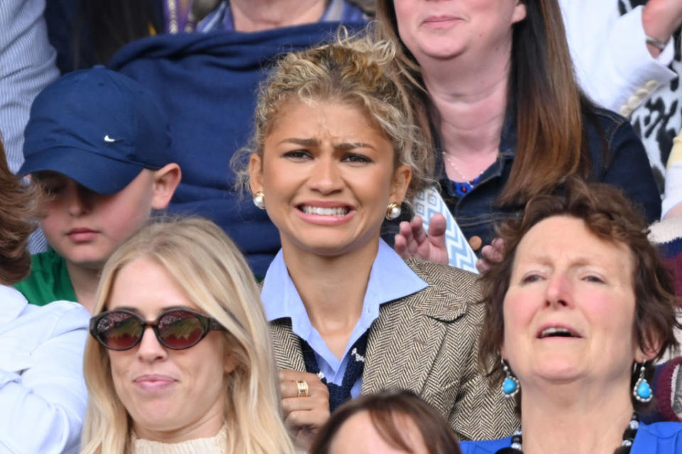 Zendaya in the middle of a crowd, wearing a formal layered outfit with a blazer. People around her display mixed reactions, appearing to be at an event