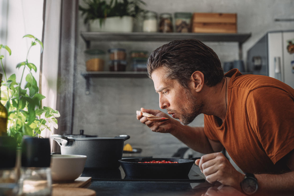 Close up shot of a handsome young happy Caucasian man tasting sauce with a mixing spoon with his eyes closed over a frying pan in a kitchen.