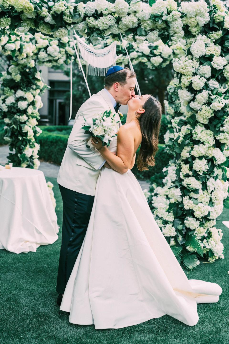 A bride and groom kiss in front of an oversized floral archway.