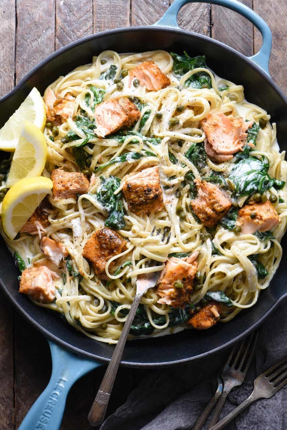 A pan of creamy pasta with chunks of salmon, spinach, and lemon wedges on a wooden surface. A fork is placed in the pan