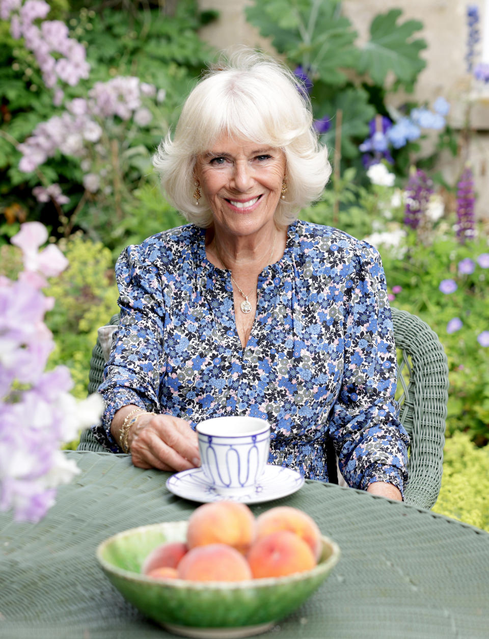 Camilla is wearing a blue floral Sophie Dundas summer dress while posing for the photograph in her garden with a cup of tea and a bowl of peaches on the table. - Credit: Clarence House via Getty Images