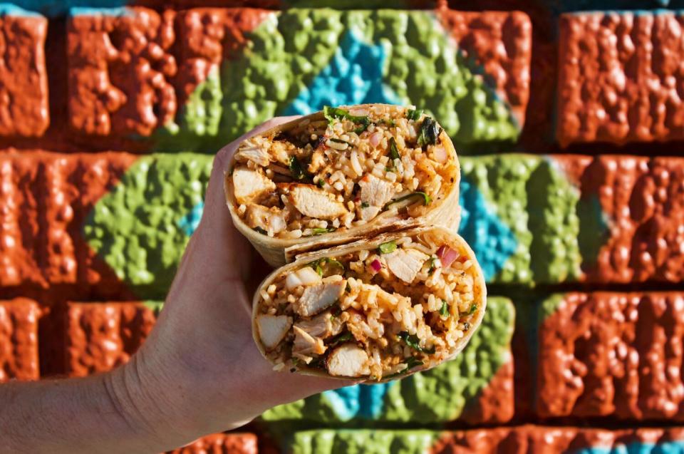 A hand holds two stacked halves of a chicken burrito in front of a mural at Mid East Tacos in Silver Lake