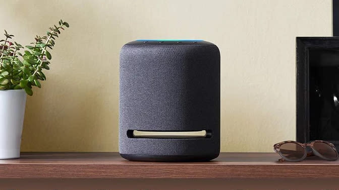 Amazon Echo Studio smart speaker sitting on a shelf next to a pot plant and sunglasses.