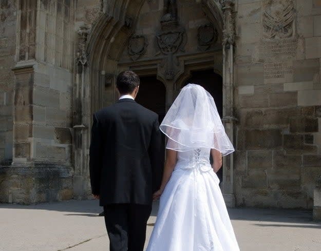 A bride and groom, both in formal wedding attire, walk hand-in-hand towards a large, ornate church entrance. Their backs are facing the camera