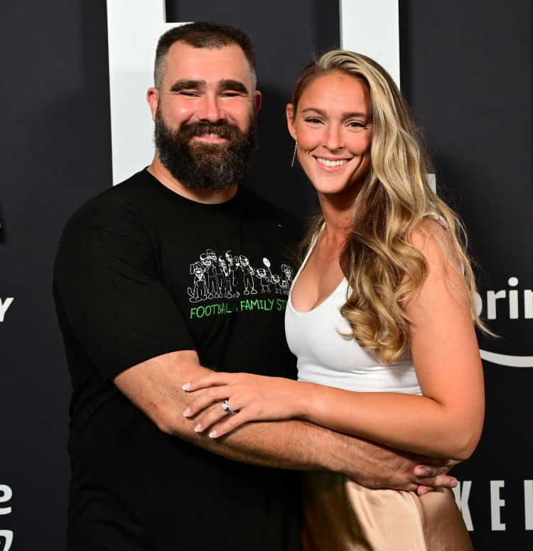 Jason Kelce and Kylie McDevitt Kelce smiling, with Jason in a casual outfit and Kylie in a stylish white top and satin skirt, at an event backdrop