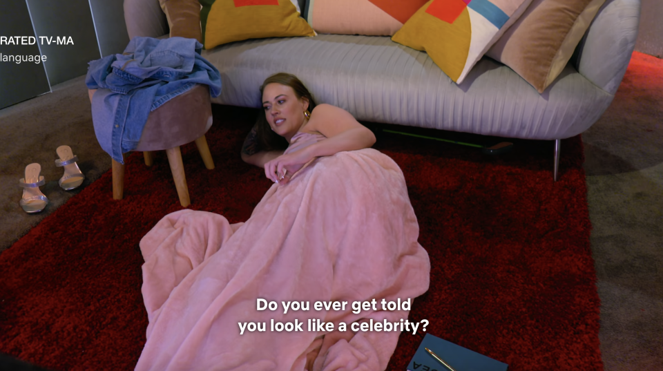 A person lounges on a couch, wearing a flowing pink outfit, with subtitle text "Do you ever get told you look like a celebrity?"