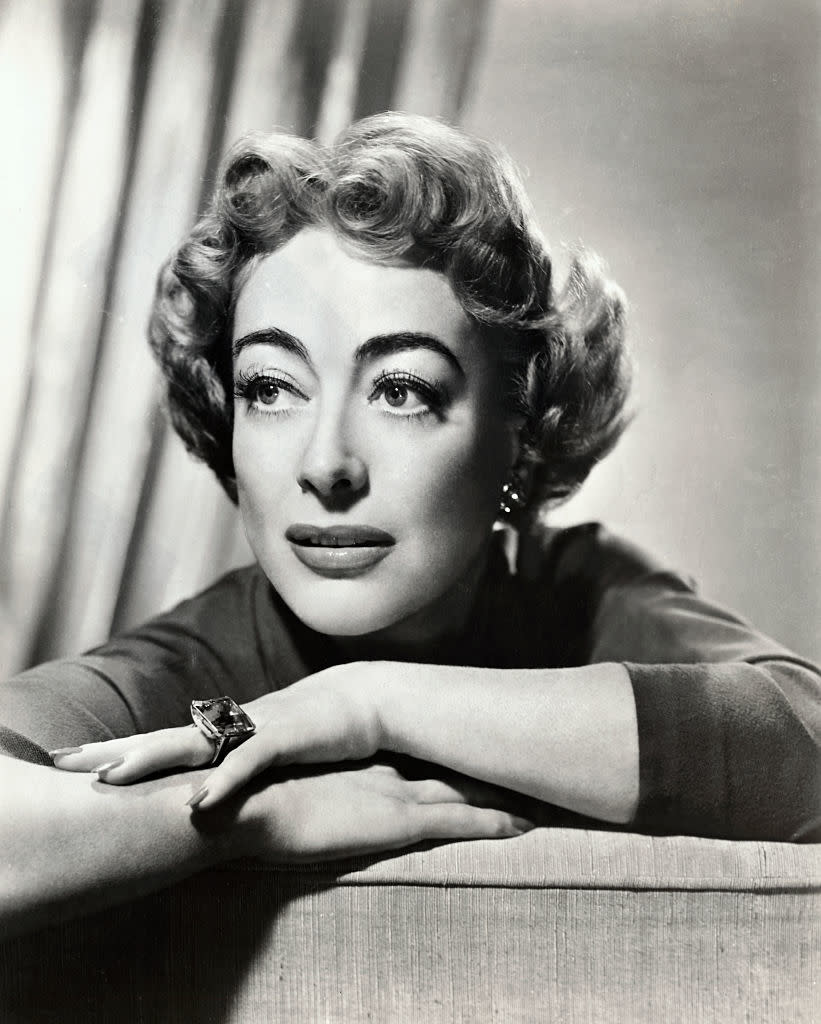 Joan Crawford resting her chin on her arms, gazing to the side with a thoughtful expression. Her short hair is styled in waves, and she wears a large ring on one hand