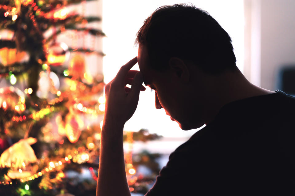 A man leans on his hand, looking pensive, as he sits in front of a lit up Christmas tree at home