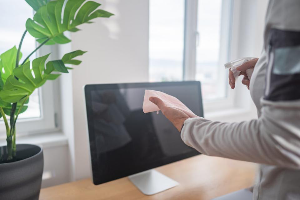 A woman spraying screen cleaner onto a microfiber cloth in front of her desktop monitor.